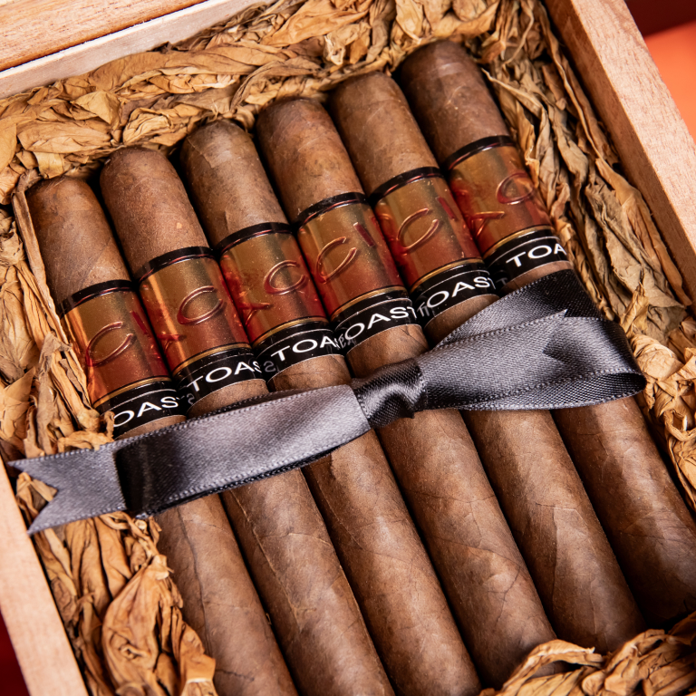 A wooden cigar box filled with six Drew Estate Acid Blue Cigars, specifically Toast Maduro cigars, nestled in shredded brown paper. Each cigar features a clear band with "TOAST" written in black, and a dark gray ribbon is tied across the ce